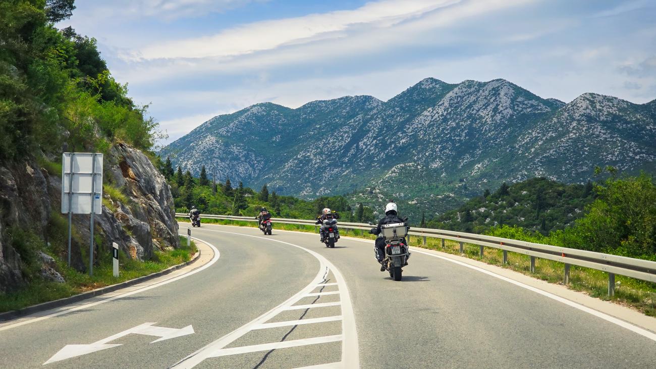 Group of motorcyclists cruising along a Croatian coastal highway with towering mountains in the background.