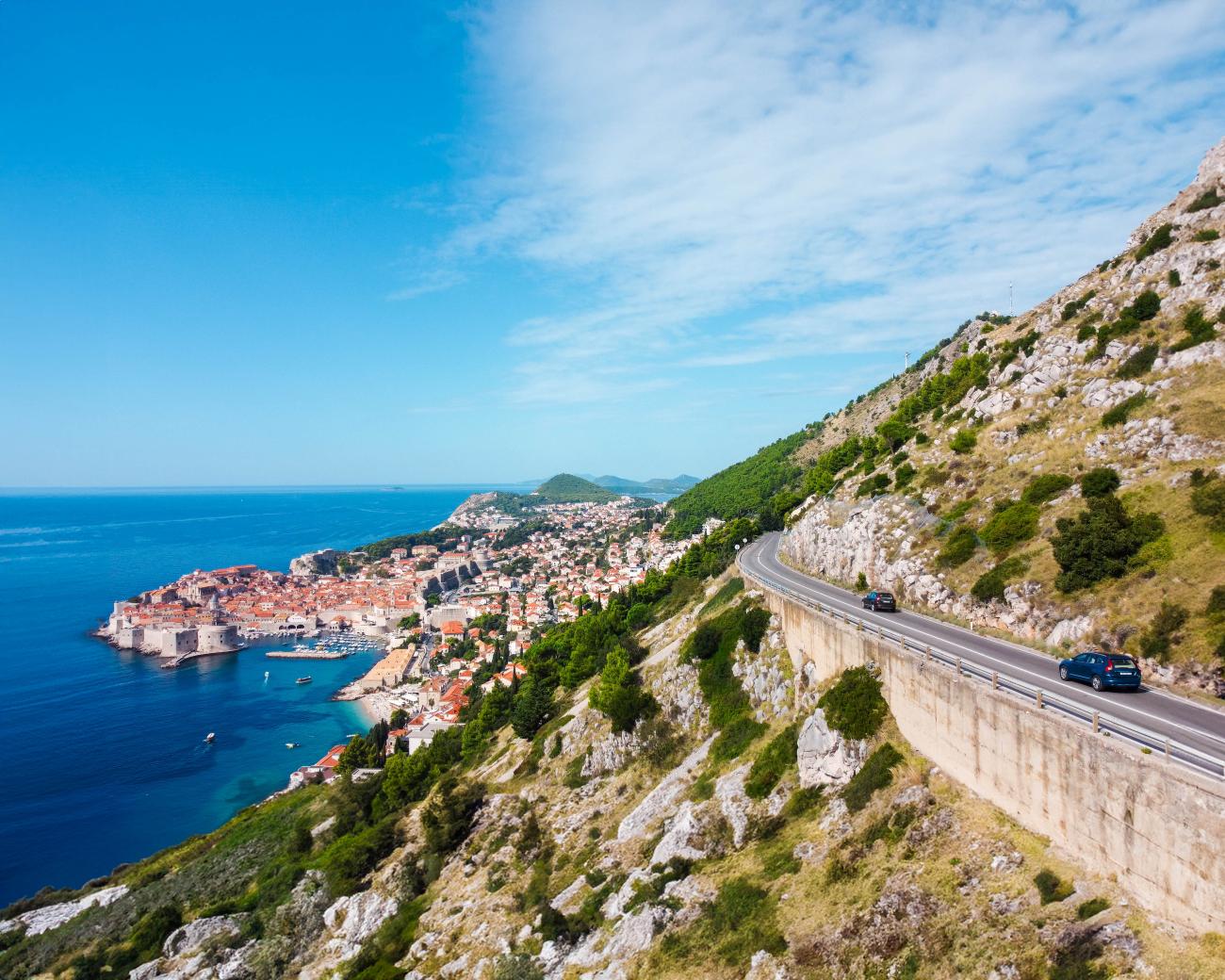 Cars driving along a cliffside coastal road above Dubrovnik Old Town with the Adriatic Sea below