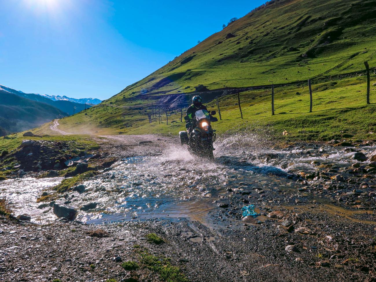 Adventure motorcyclist splashing through a mountain stream crossing on a dirt trail in the green Caucasus hills.