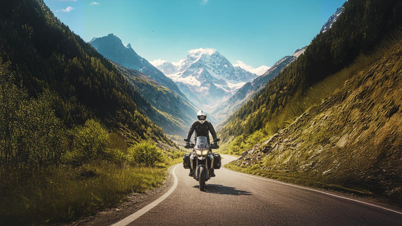 Motorcyclist riding through a lush green Caucasus mountain valley with snow-capped peaks ahead on a winding Georgian road.
