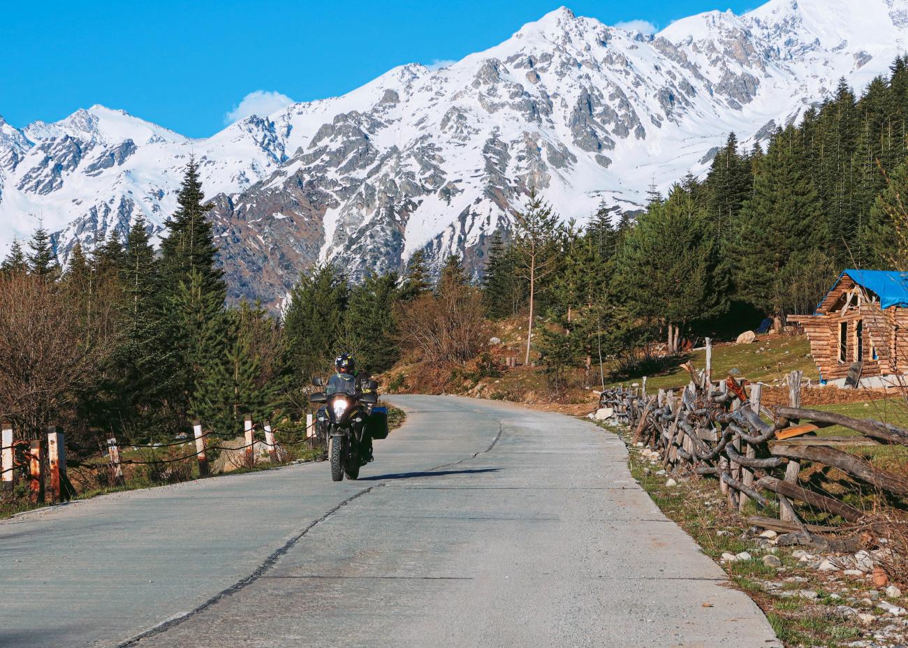 Motorcyclist riding along a mountain road in Svaneti with dramatic snow-covered Caucasus peaks and pine forests.
