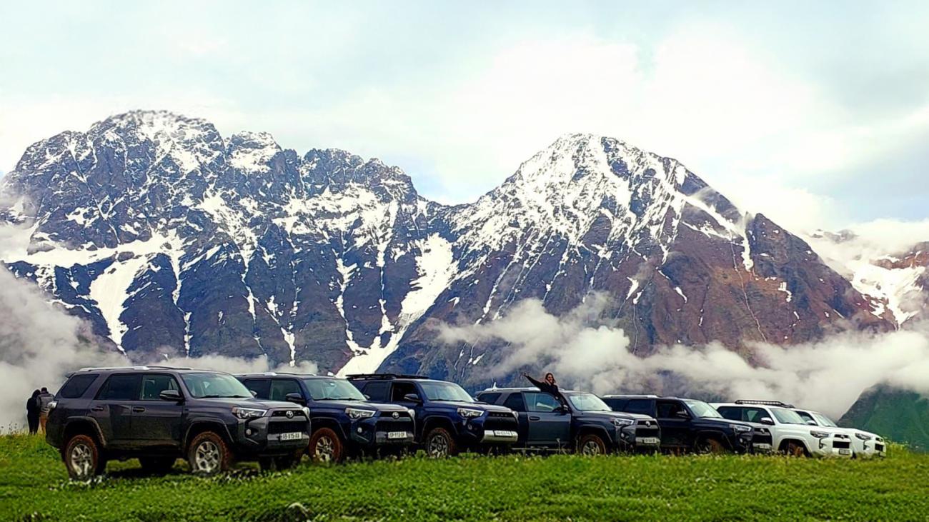 A fleet of Toyota 4Runner SUVs lined up on a green alpine plateau with towering snow-covered Caucasus peaks and clouds behind them