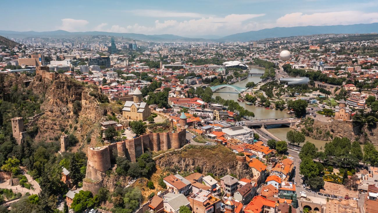 Aerial panorama of Tbilisi showing the ancient Narikala Fortress walls on a cliff above the old town with the modern city skyline beyond