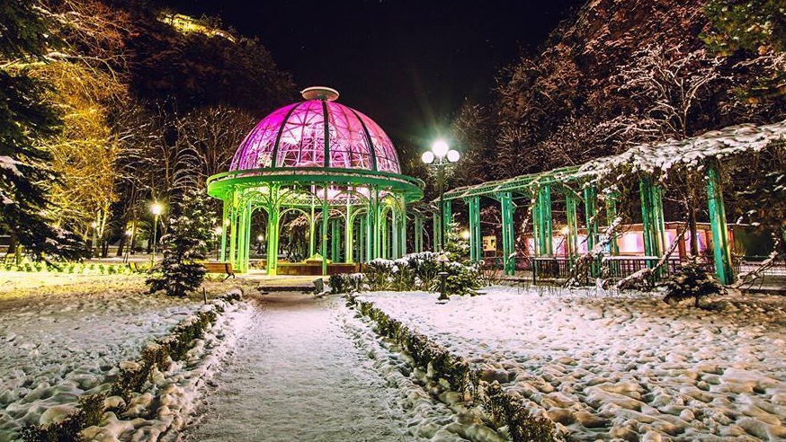 A colorfully illuminated glass gazebo in Borjomi Central Park surrounded by snow-covered trees on a winter night