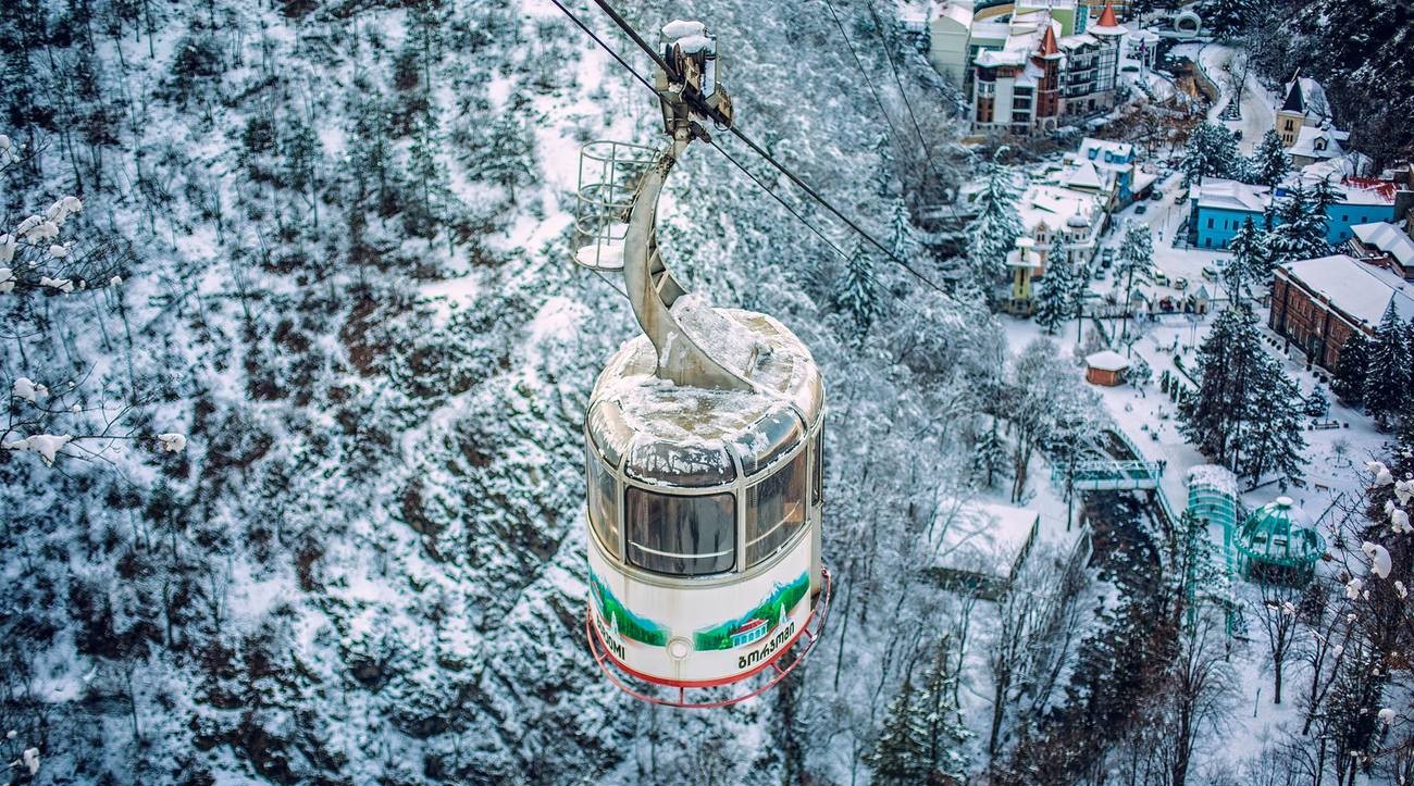 An aerial cable car gondola glides over a snow-blanketed forested valley and mountain town in Georgia
