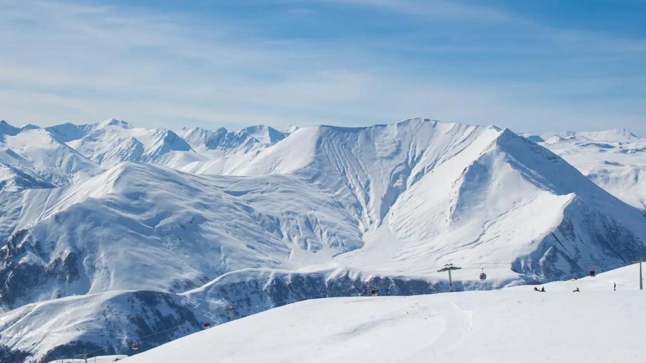 A panoramic view of dramatic snow-covered Caucasus mountain peaks with ski lifts visible on the slopes below