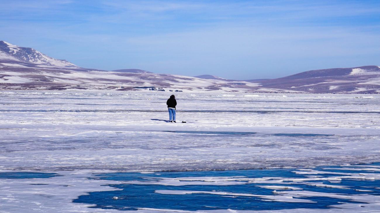 A lone person stands on a frozen lake with cracked blue ice stretching toward snow-dusted hills under a clear winter sky
