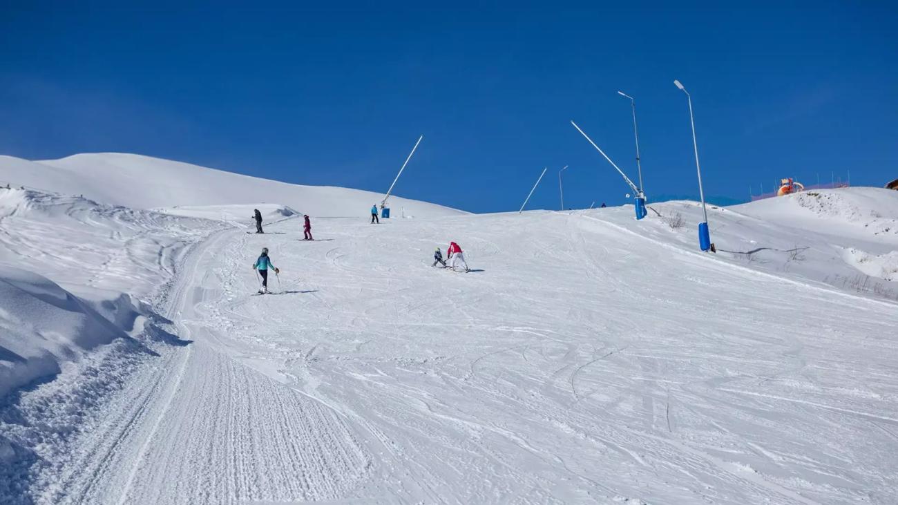 Skiers descend a sunny groomed slope at a Georgian ski resort with pristine snow and blue skies