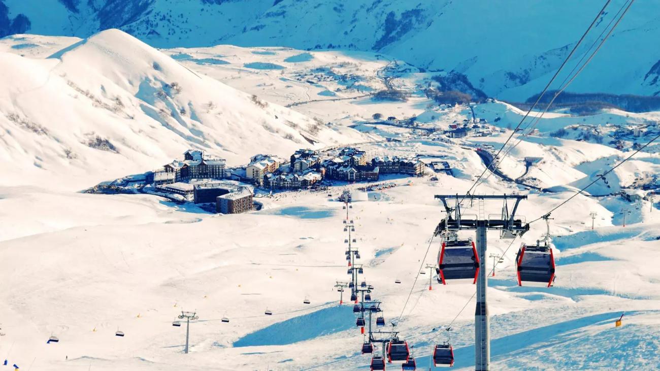 Red gondola cable cars ascending over a sprawling snow-covered ski resort with the Caucasus mountains and resort buildings in the background