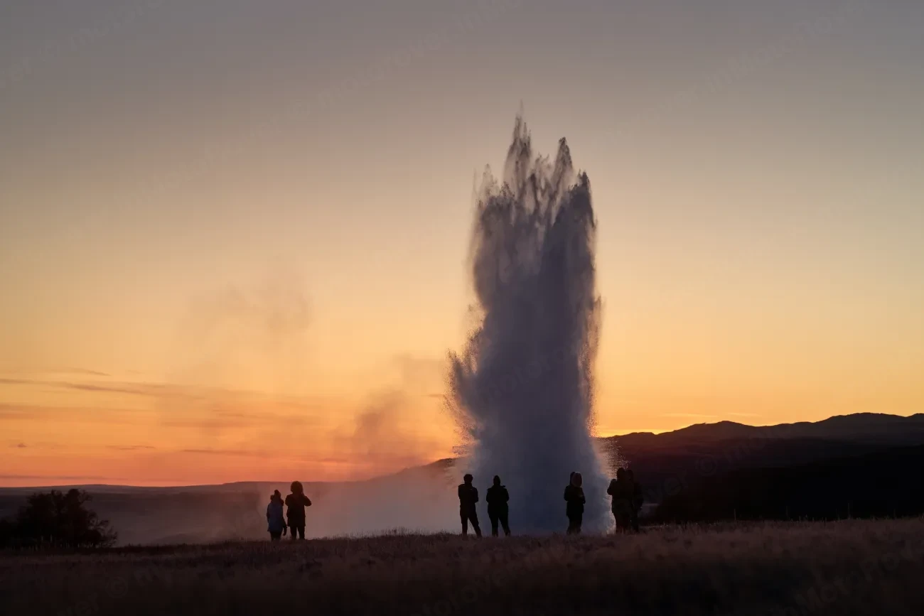 A geyser erupting against an orange sunset sky, watched by visitors