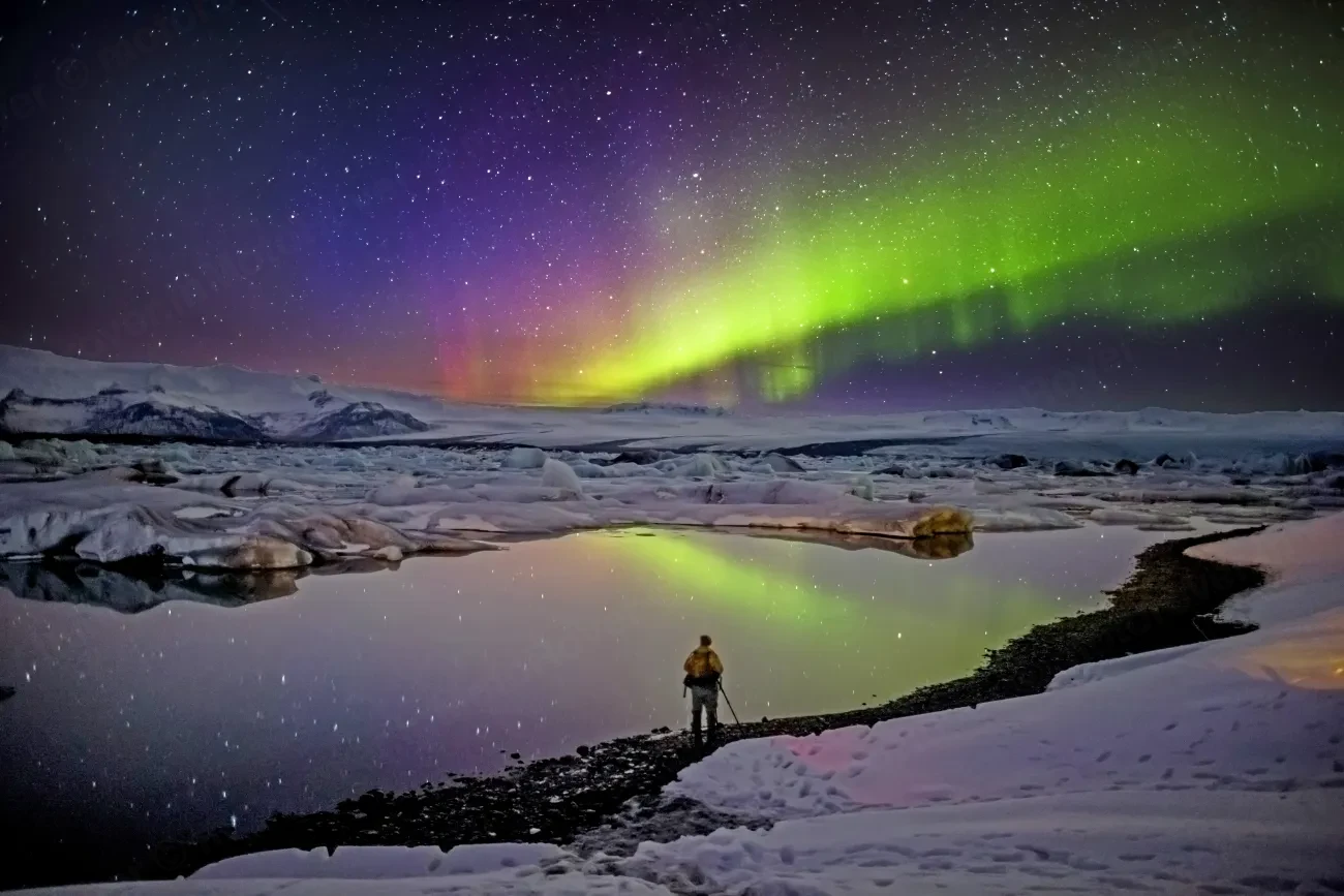 Green and violet northern lights over an icy lagoon in Iceland, with a photographer on the shore