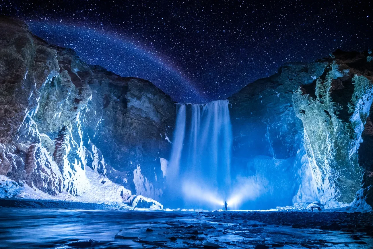 A waterfall pouring between snow-covered cliffs under a starry winter night sky