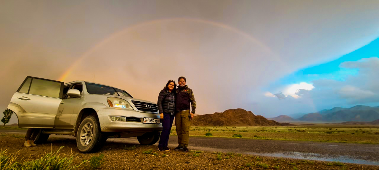 A couple poses beside their SUV under a full rainbow arching over the Kyrgyz mountain landscape at sunset.