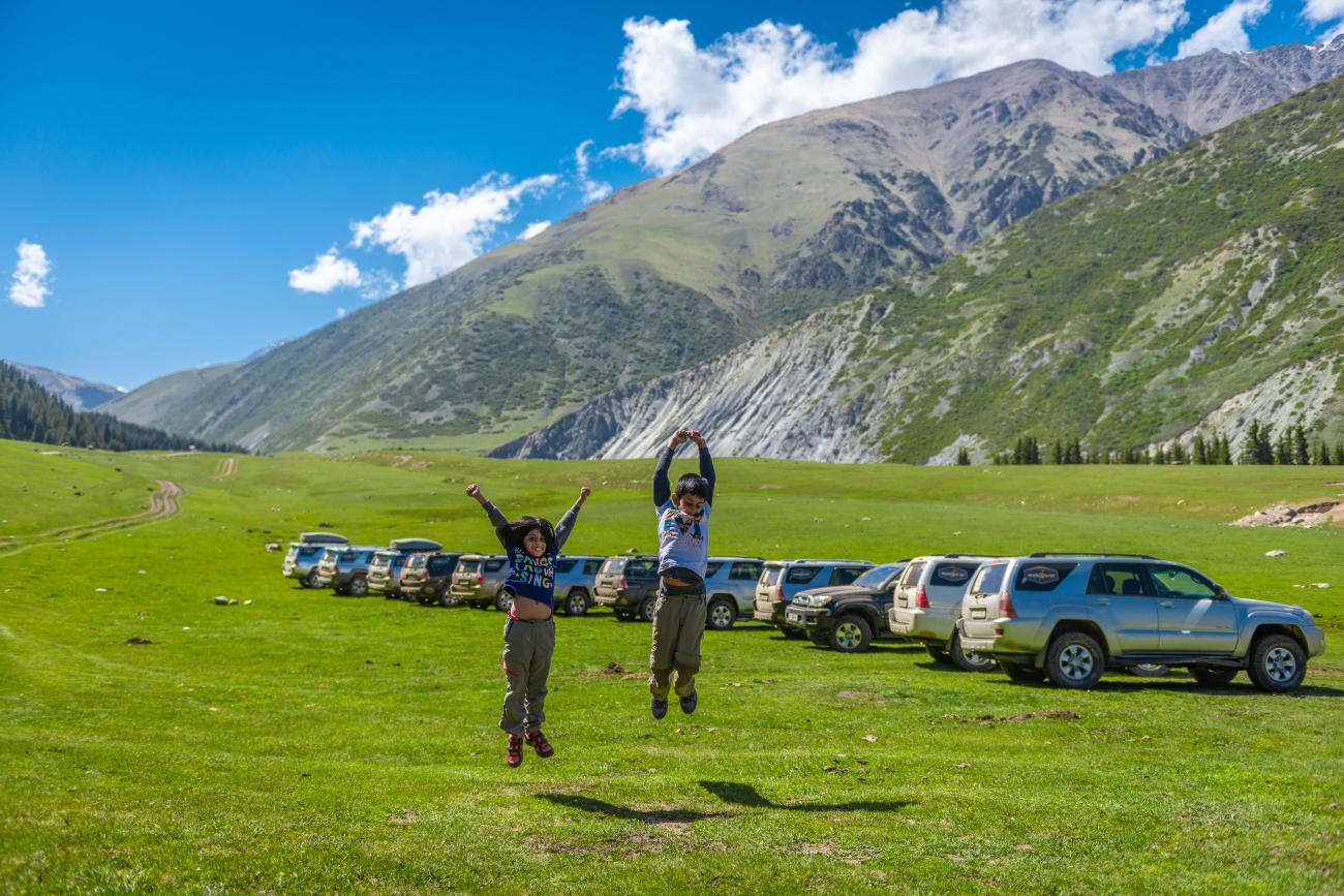 Two travellers jumping for joy on a green alpine meadow surrounded by parked SUVs and towering Kyrgyz mountains.