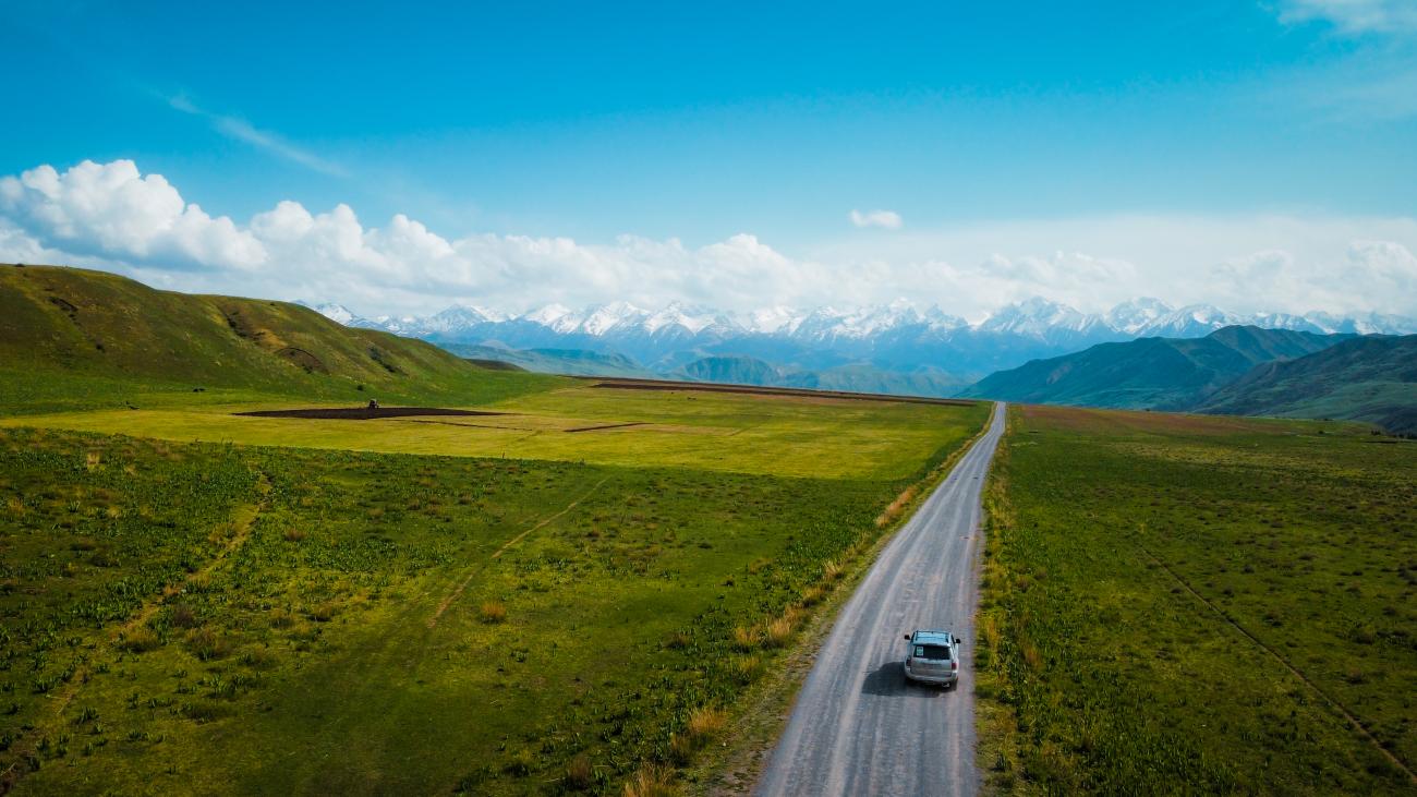 A lone SUV drives along an open road through green spring pastures toward a wall of snow-capped peaks in Kyrgyzstan.