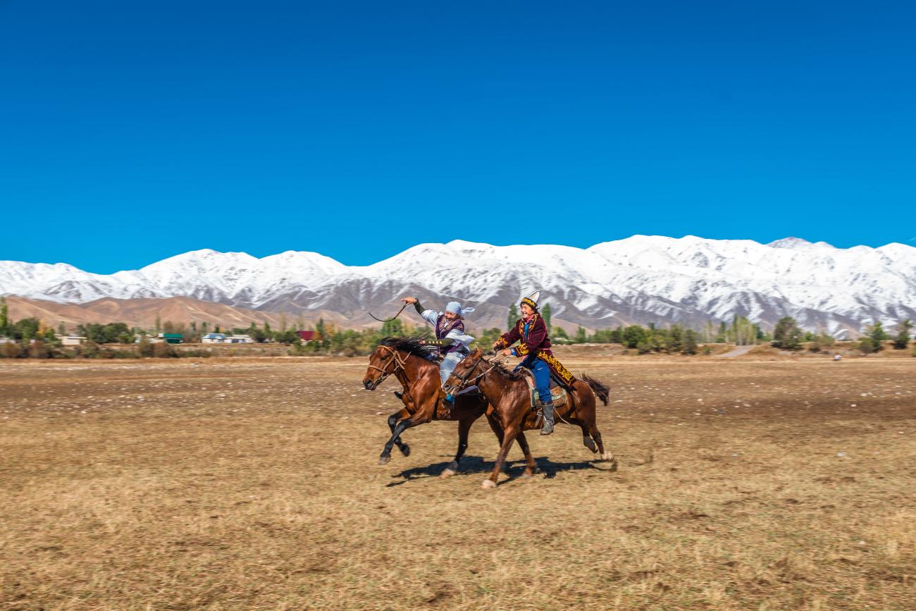 Two Kyrgyz riders in traditional dress galloping on horseback across a plain with snowy mountains in the distance.