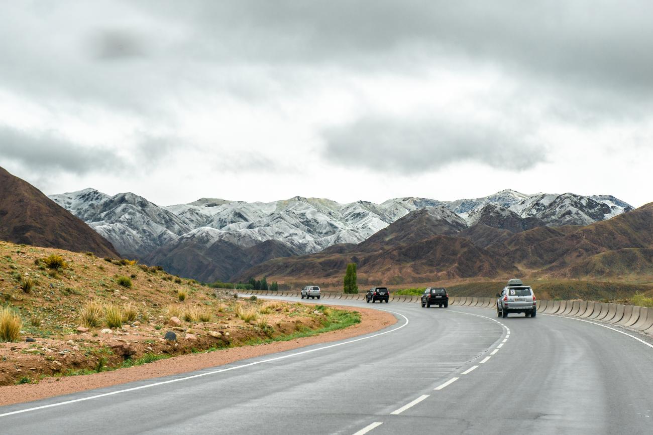 SUVs drive along a sweeping paved highway curving toward snow-dusted mountains in Kyrgyzstan under overcast skies.