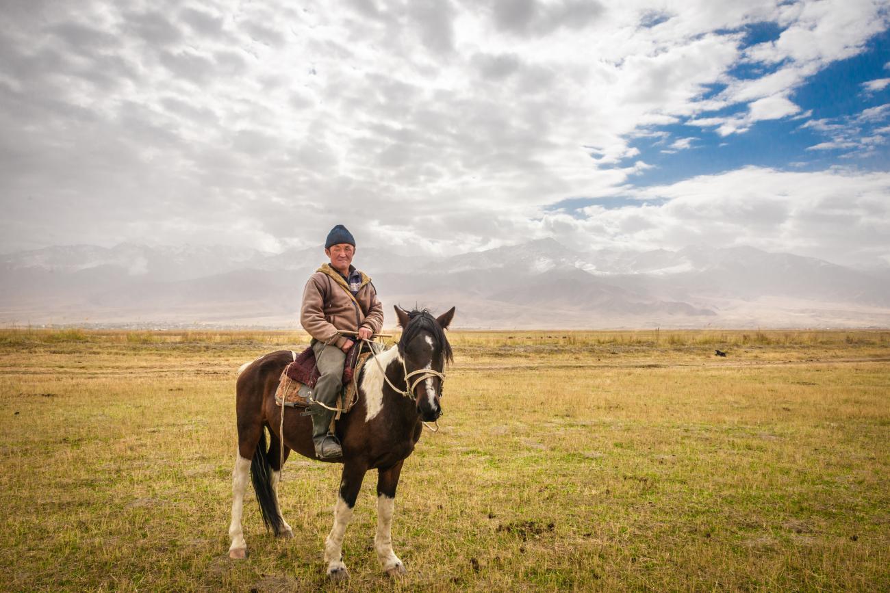 A Kyrgyz horseman sits on his horse in the middle of a vast golden steppe with hazy mountains stretching across the horizon.