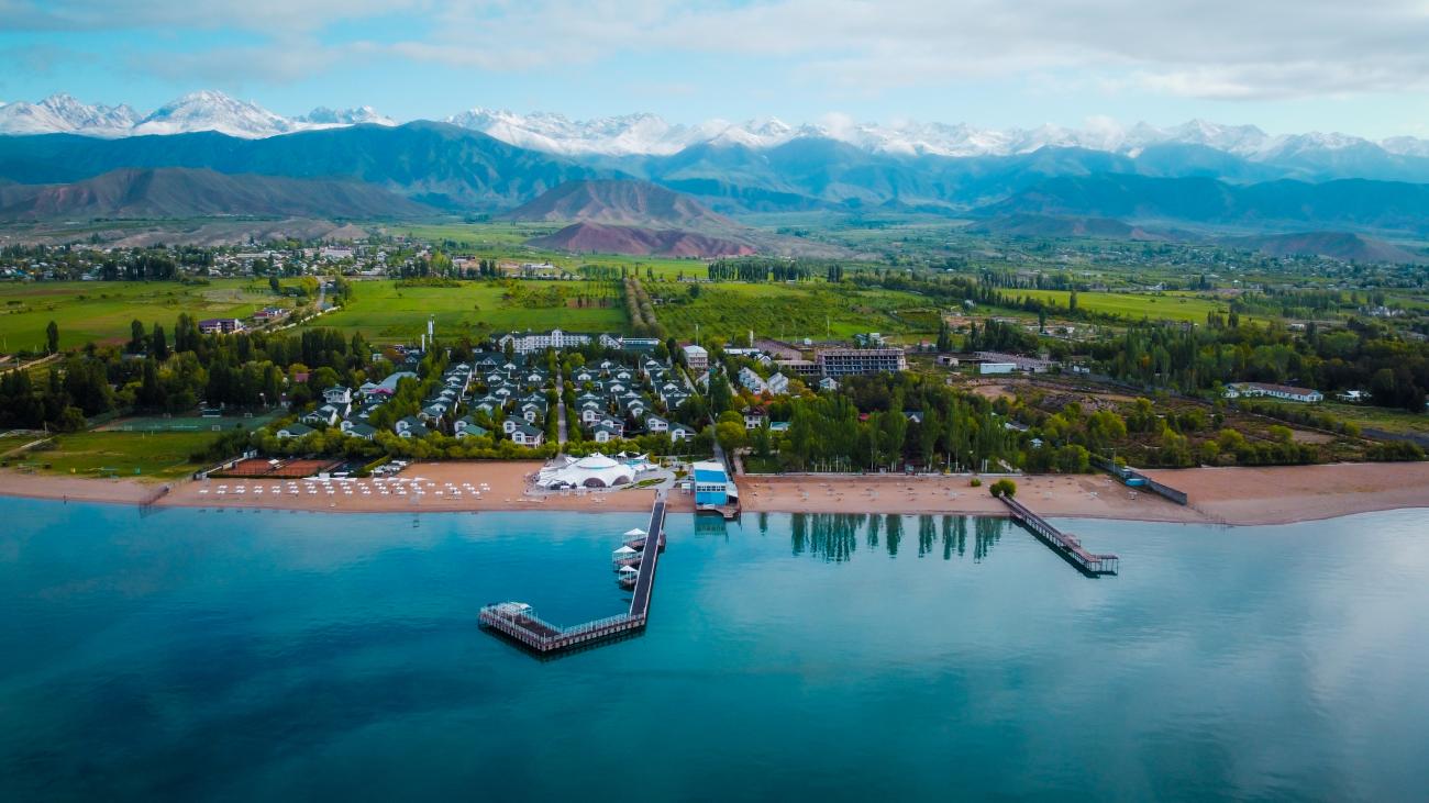Aerial view of a lakeside resort on the shore of Issyk-Kul with a pier extending into turquoise water and snow-capped Tien Shan mountains beyond.