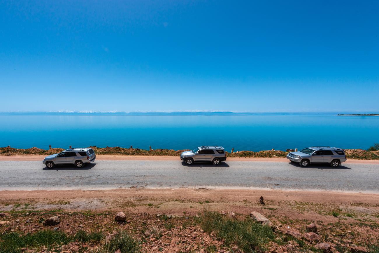 SUVs parked along the shore road beside the turquoise waters of Issyk-Kul Lake in Kyrgyzstan under a clear blue sky.