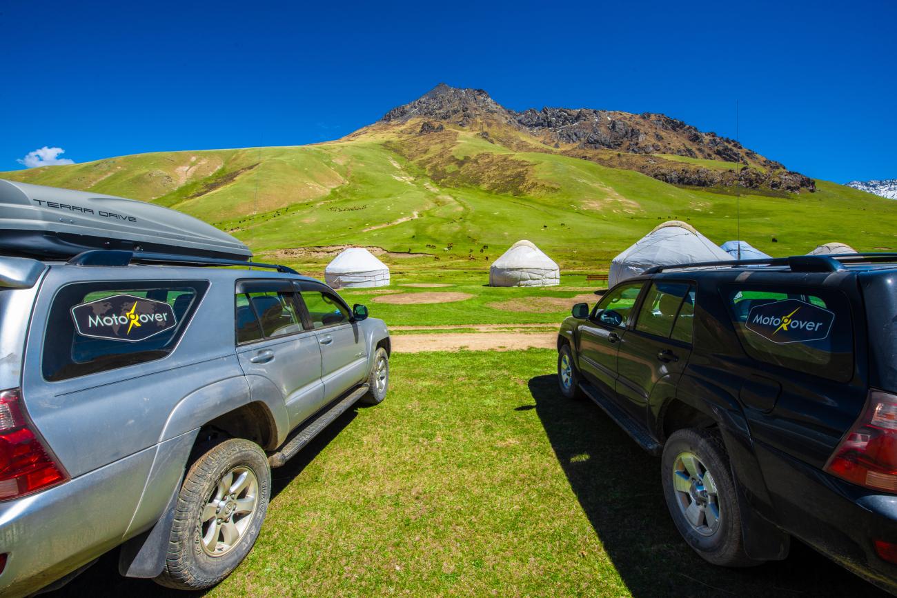 Two MotoRover-branded SUVs parked on green grass facing white yurts at the base of a rocky Kyrgyz mountain.