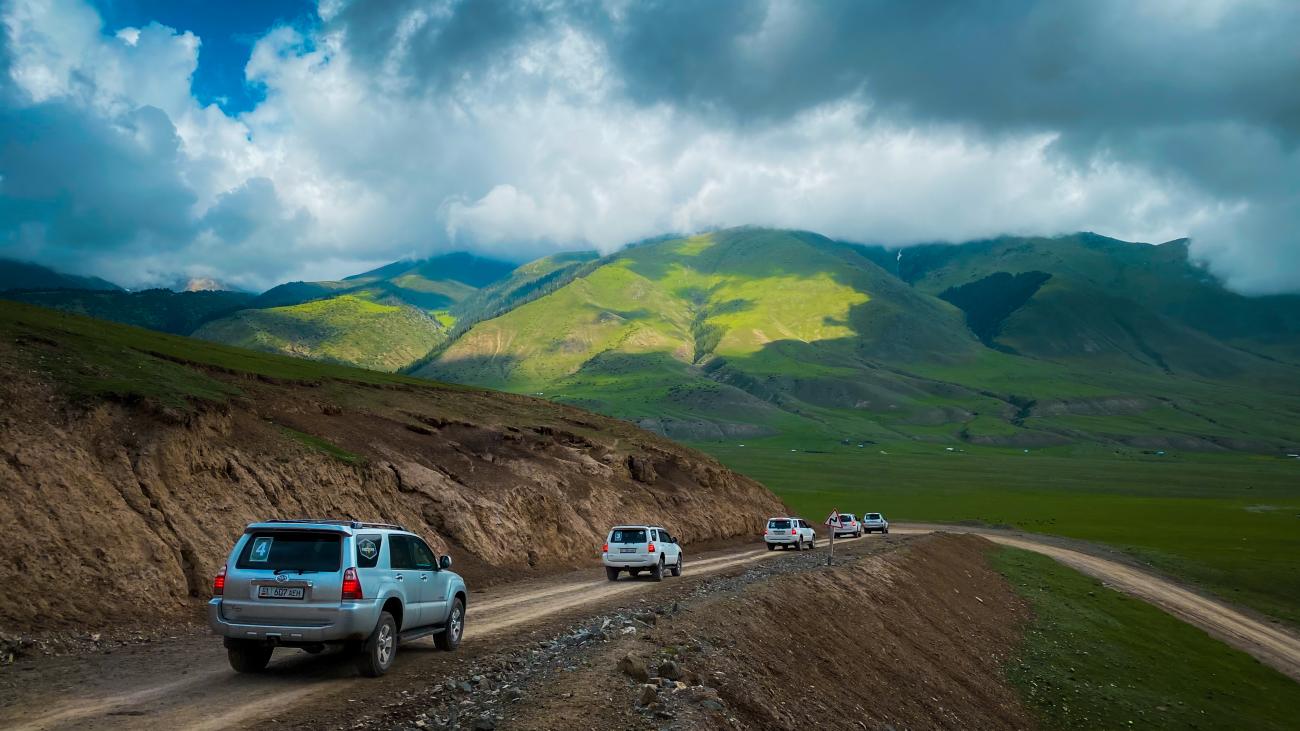 SUV convoy navigating a rugged dirt road cut into a hillside with sunlit green mountains and dramatic clouds ahead.