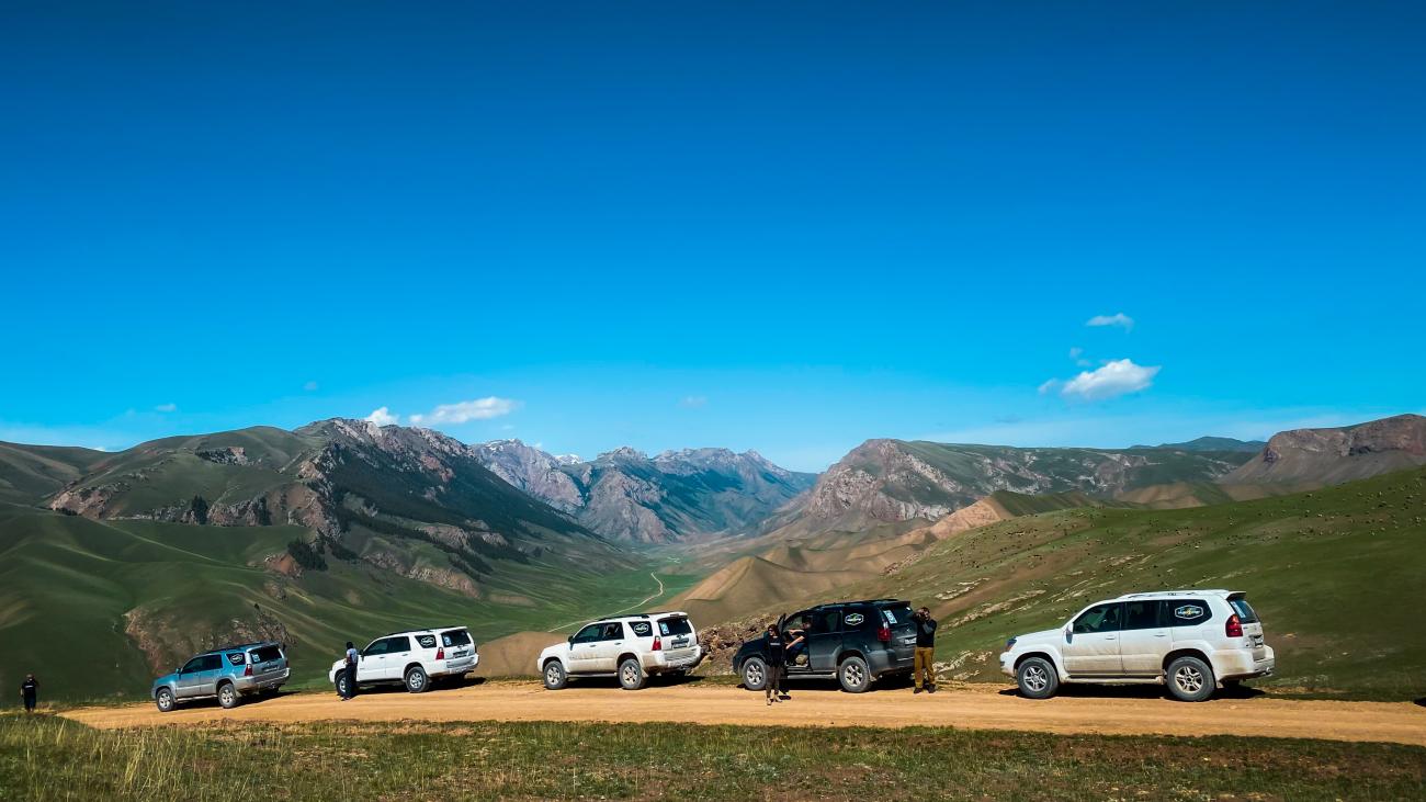 SUVs stopped on a high ridge overlooking a sweeping panorama of deep valleys and rugged Kyrgyz mountain ranges under blue sky.