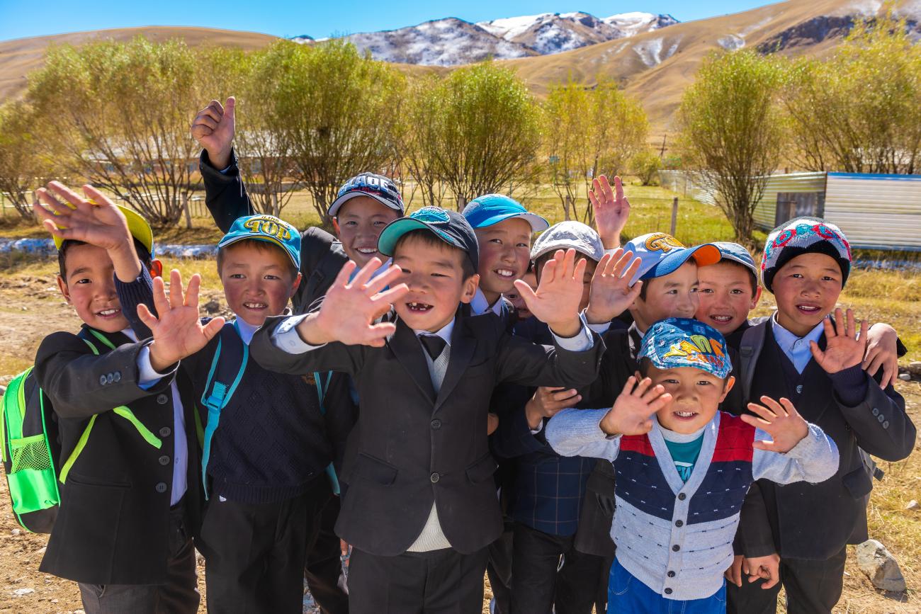 A group of smiling Kyrgyz schoolchildren wave at the camera in a mountain village with snowy peaks behind them.