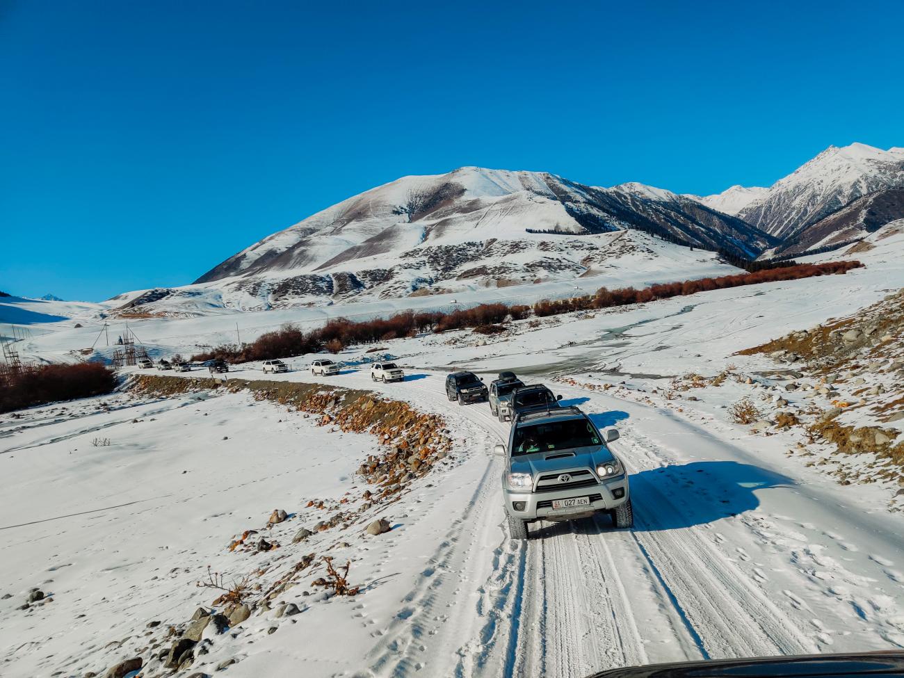 A convoy of SUVs driving through a snowy road near a mountain village with peaks towering behind