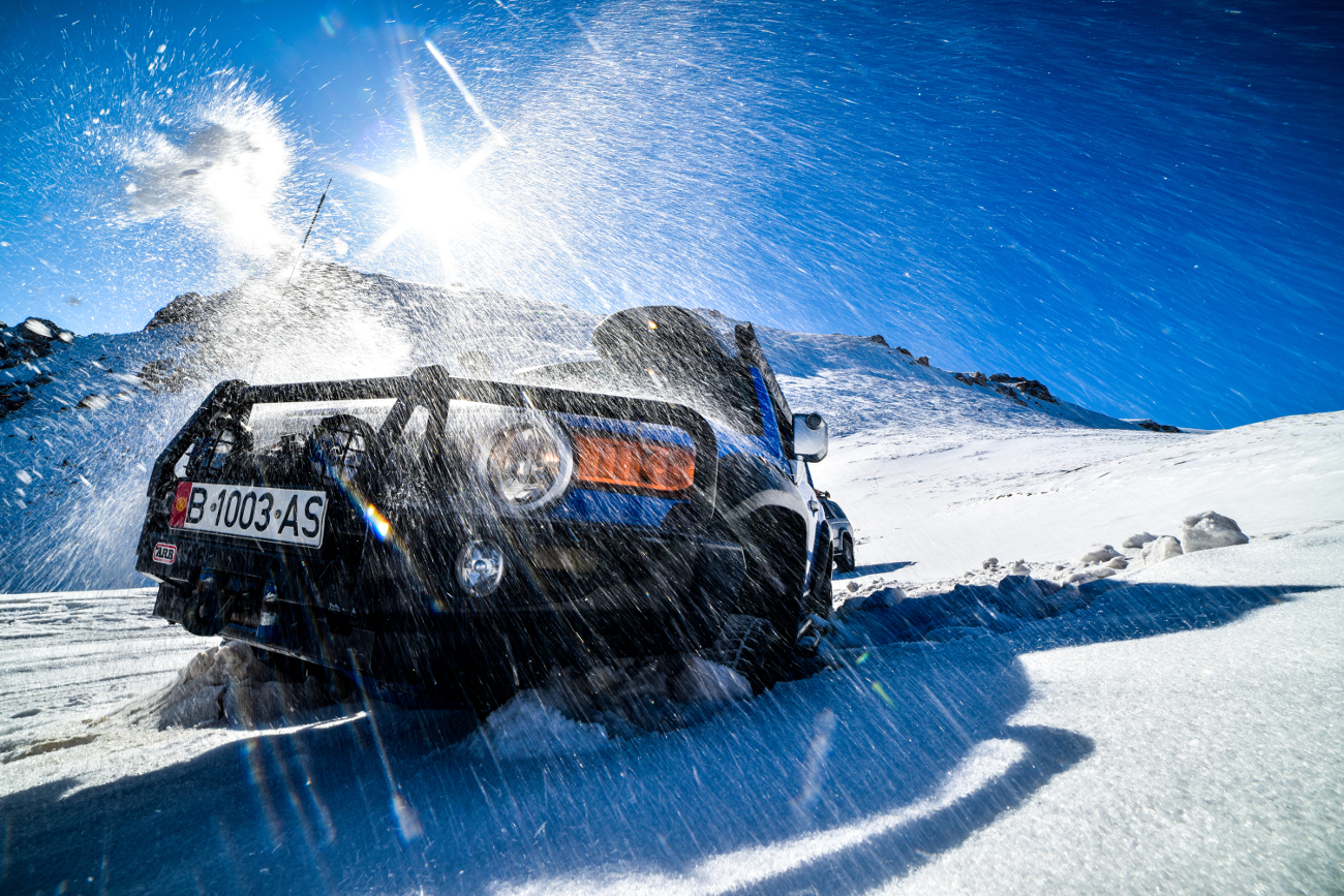 An SUV blasting through deep powder snow under bright sunlight in the Kyrgyz mountains