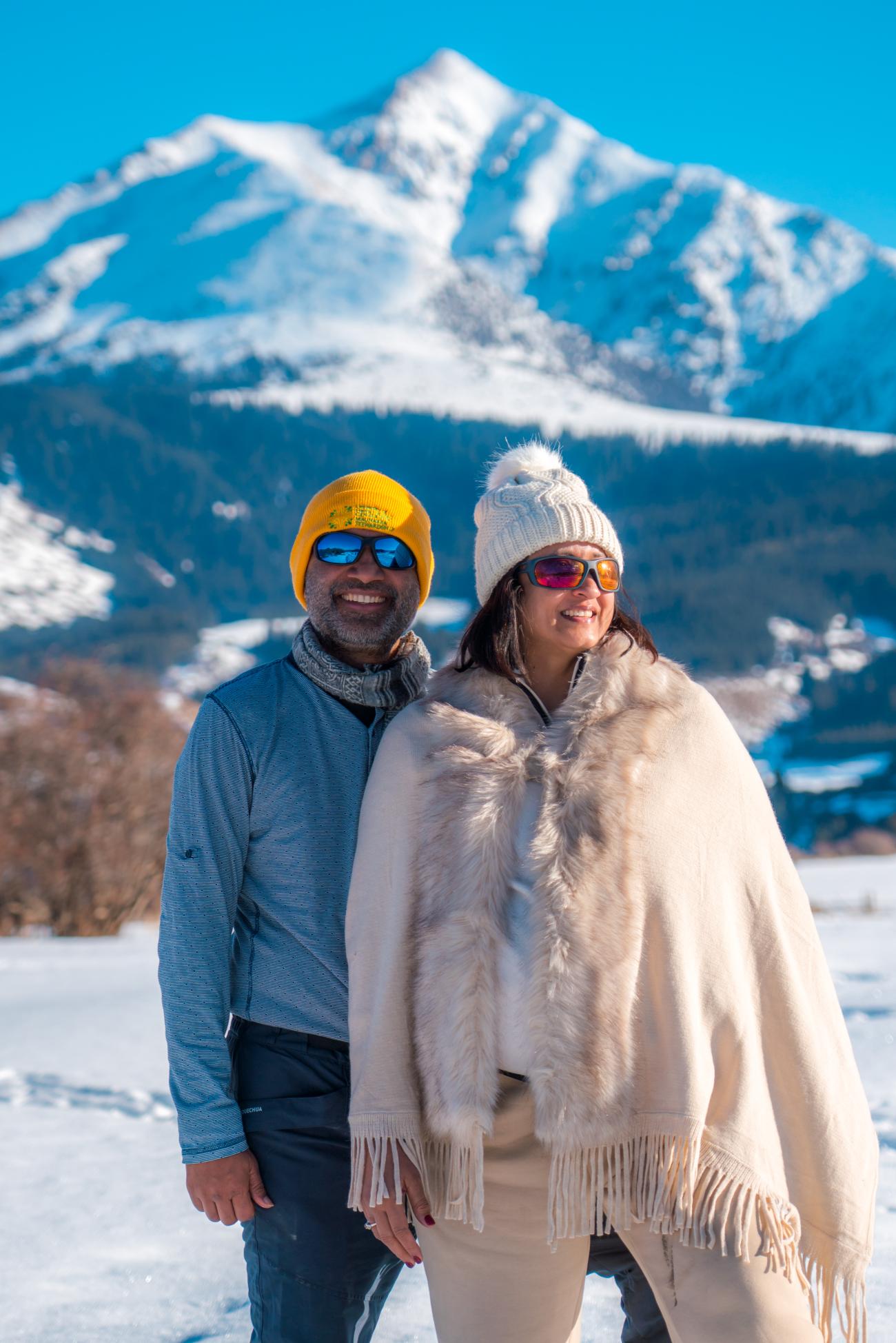 A smiling couple posing in winter gear with a dramatic snow-capped mountain peak behind them