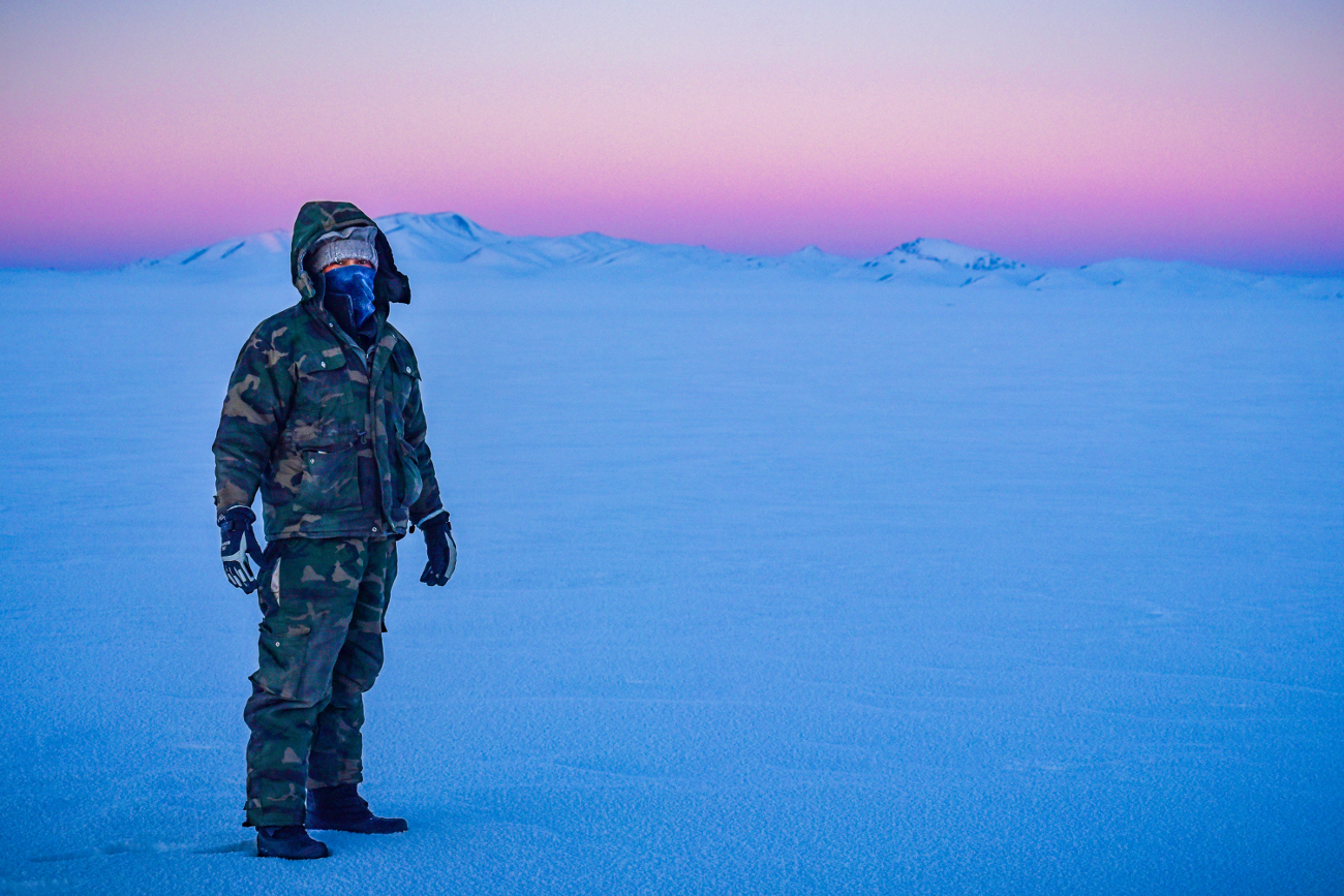 A bundled-up explorer standing on a frozen expanse at twilight with pink and blue skies over distant mountains
