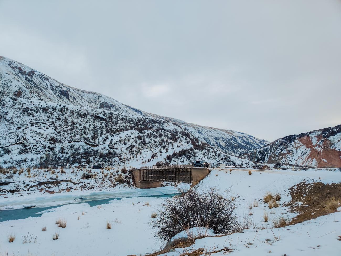A bridge over a frozen turquoise river surrounded by snow-covered mountains in Kyrgyzstan