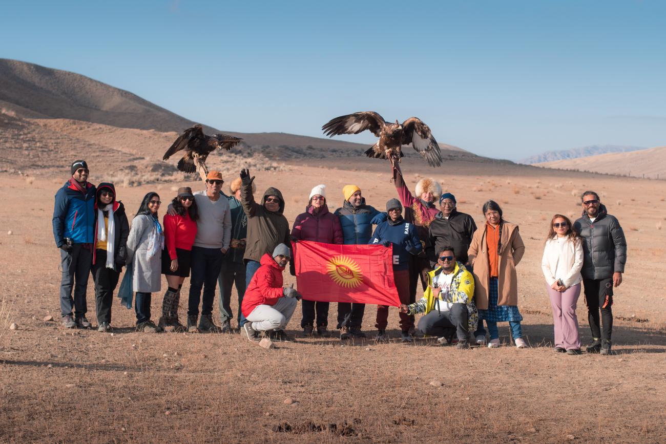 Tour group posing with eagle hunters and the Kyrgyz flag on an open steppe with mountains behind