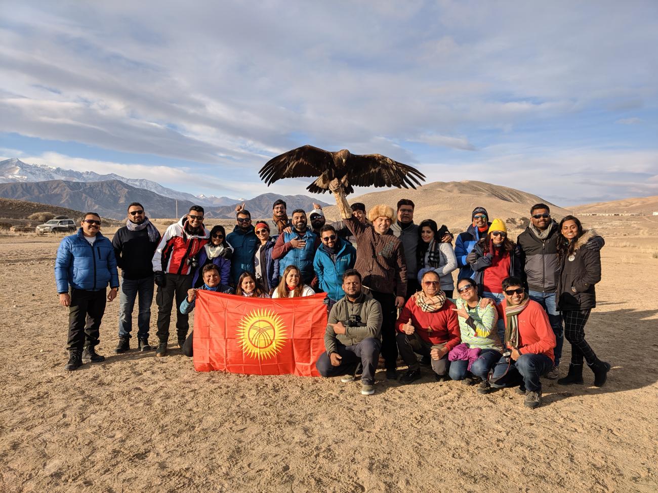 Tour group posing with Kyrgyz eagle hunters and the national flag against a mountain backdrop