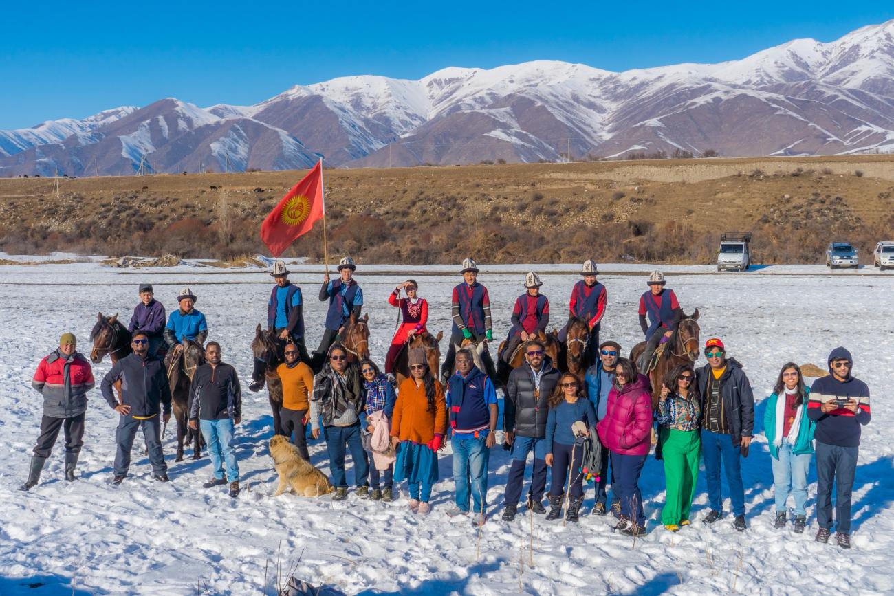 Tour group and Kyrgyz horsemen posing together with the national flag on a snowy field with mountains behind