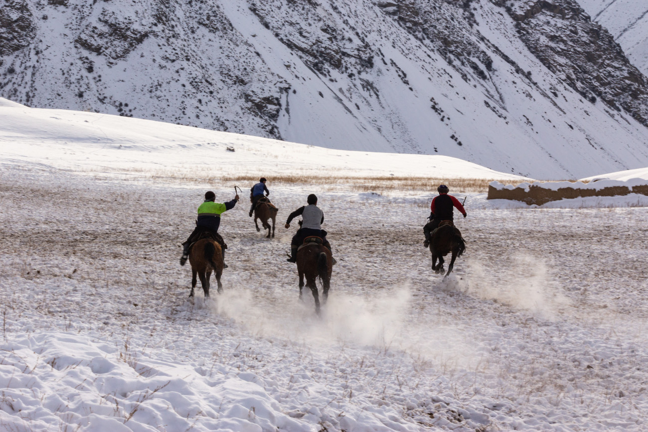Riders galloping on horseback across a snowy valley with towering mountains behind them