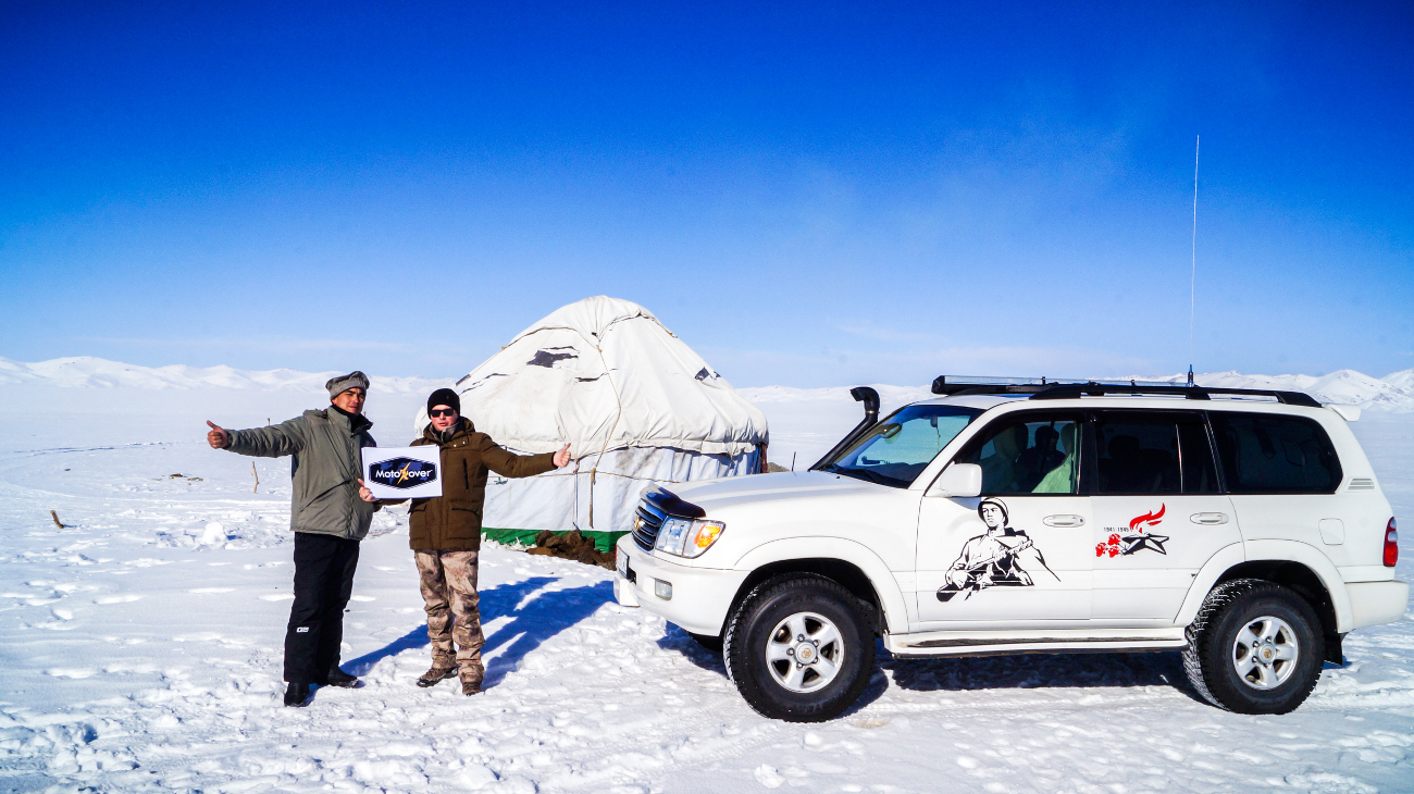 Two travelers posing with a MotoRover sign beside a snow-covered yurt and their 4x4 vehicle