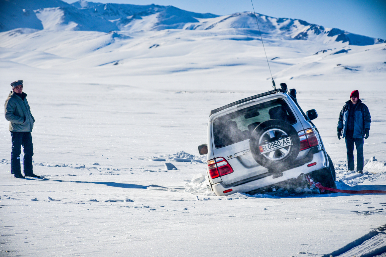 An SUV tilting into a crack on a frozen snow-covered surface while crew members assist with recovery