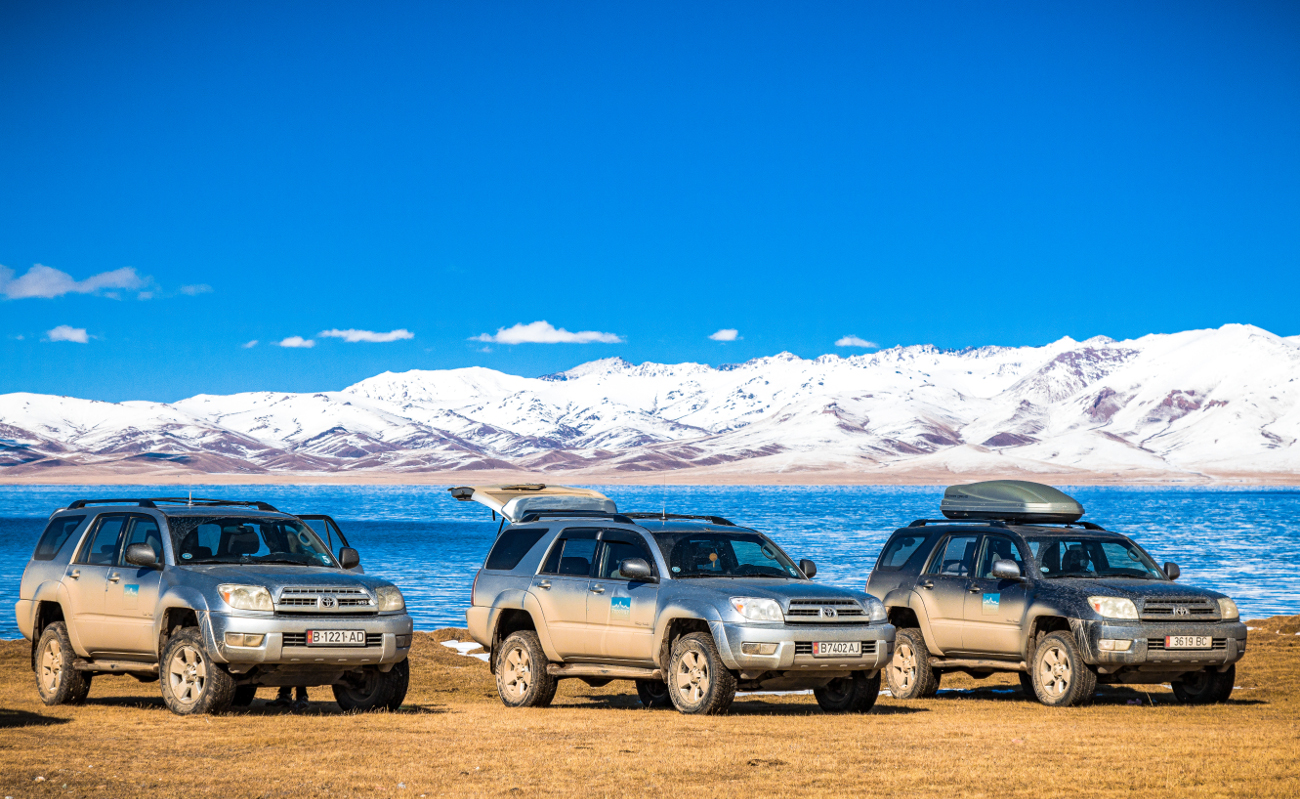 Three Toyota 4x4 vehicles parked by a vivid blue alpine lake with a panorama of snowy mountains