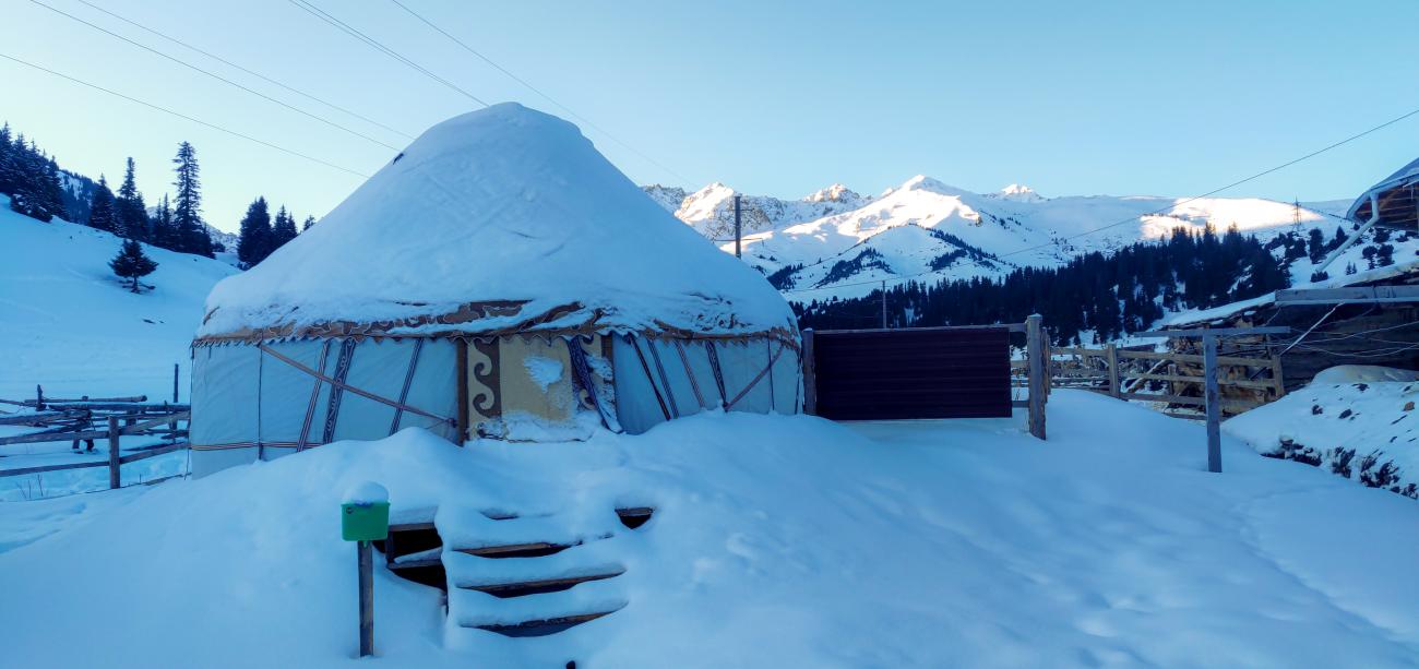 A traditional Kyrgyz yurt blanketed in snow with sunlit mountain peaks in the background