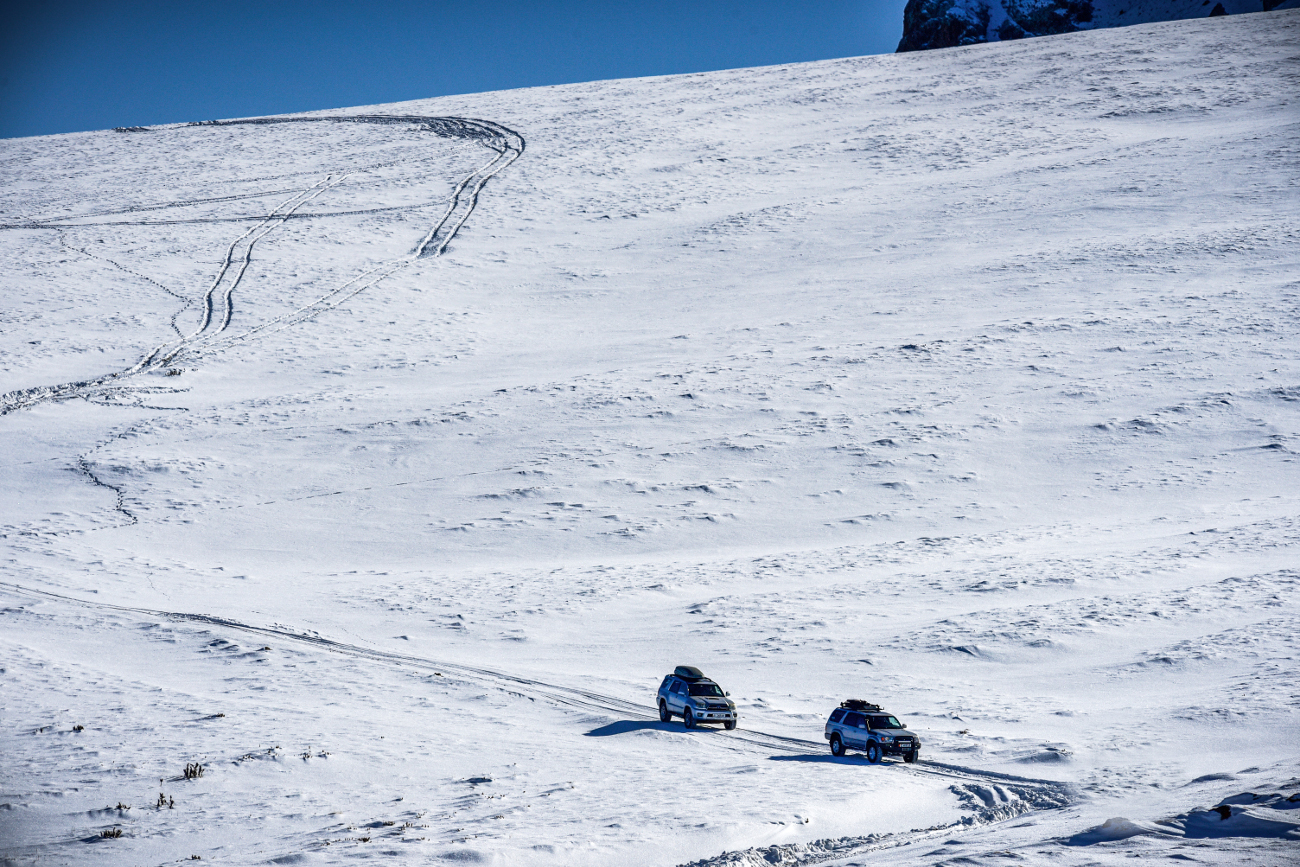 Two 4x4 vehicles crossing a vast white snow-covered plateau under a clear blue sky