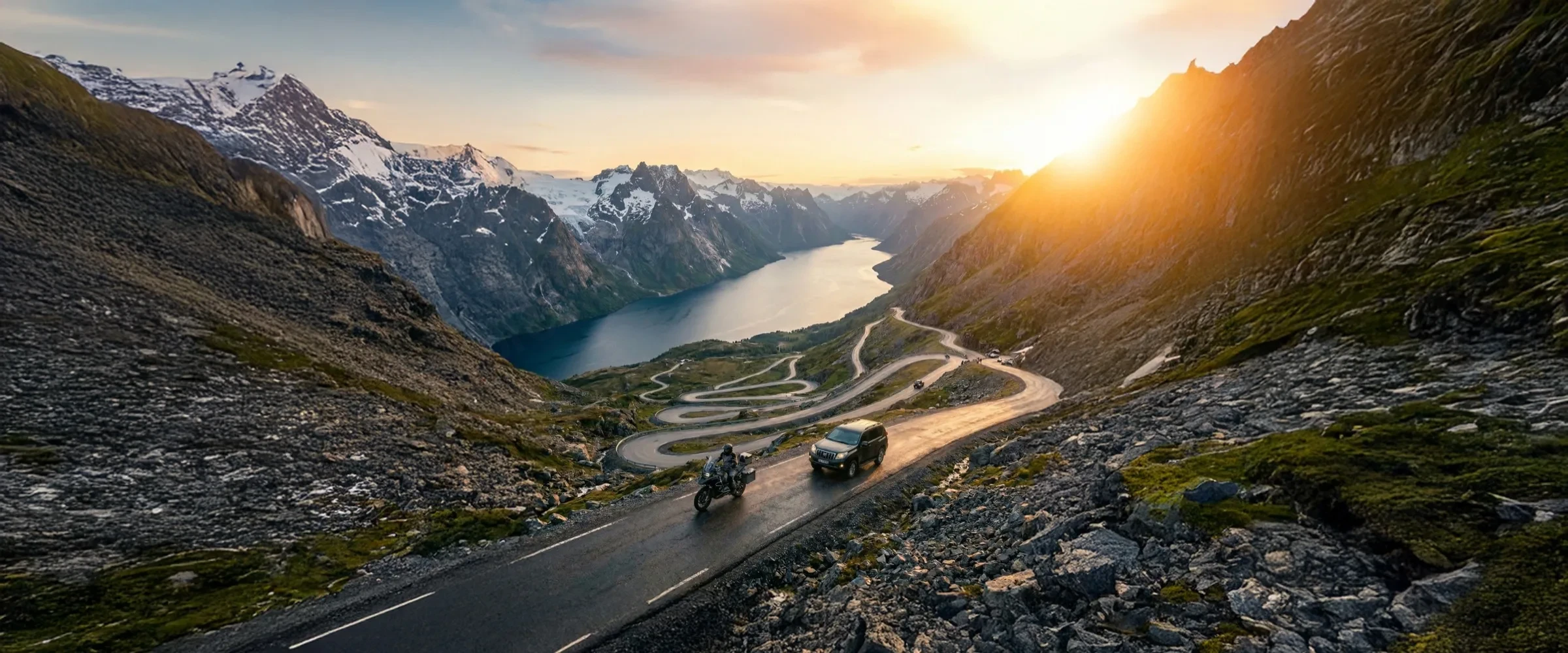 Motorcycle and support car on a fjord road framed by snow-capped peaks in Norway