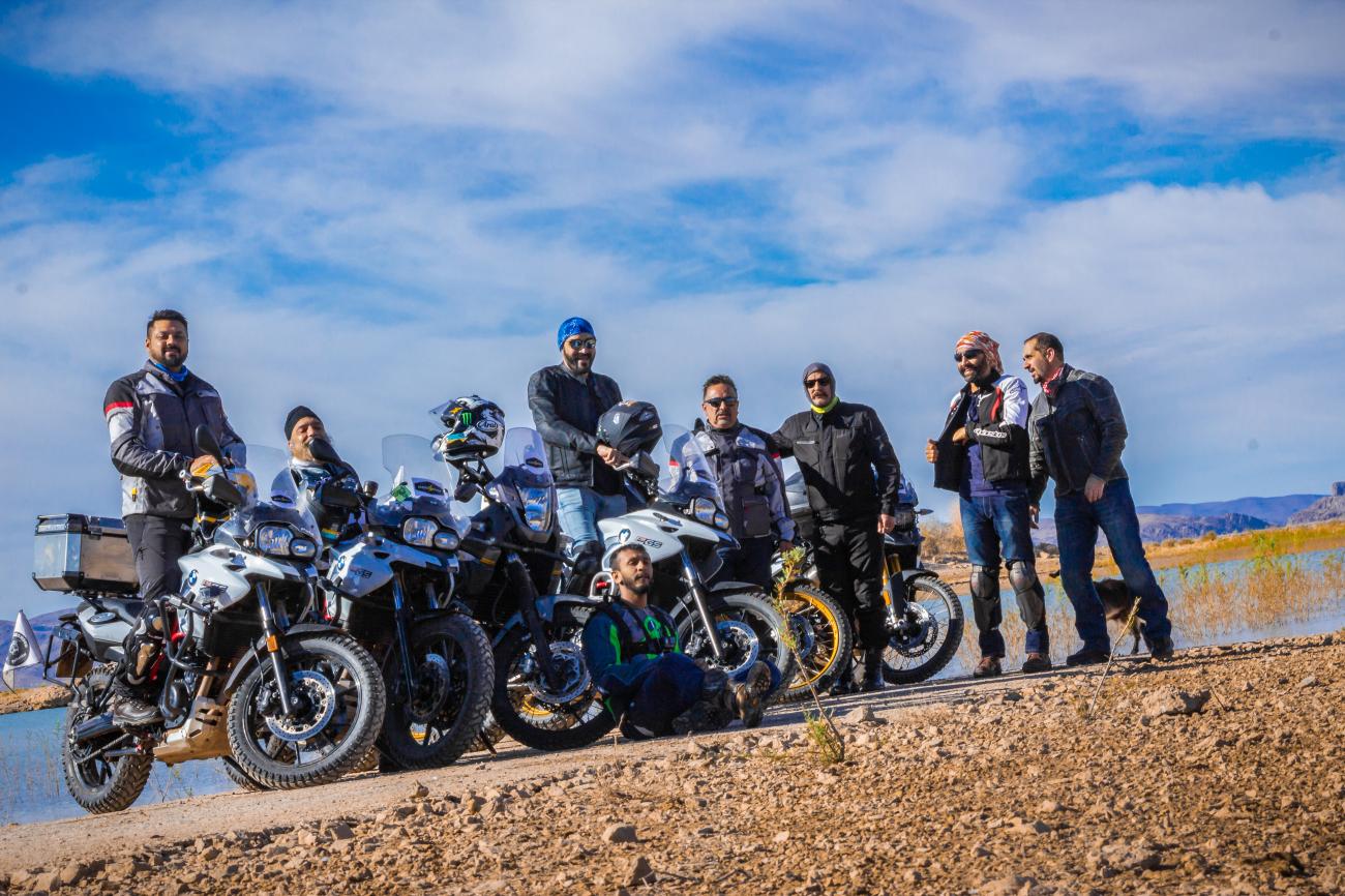 A group of riders posing with their adventure motorcycles beside a lake in Morocco.
