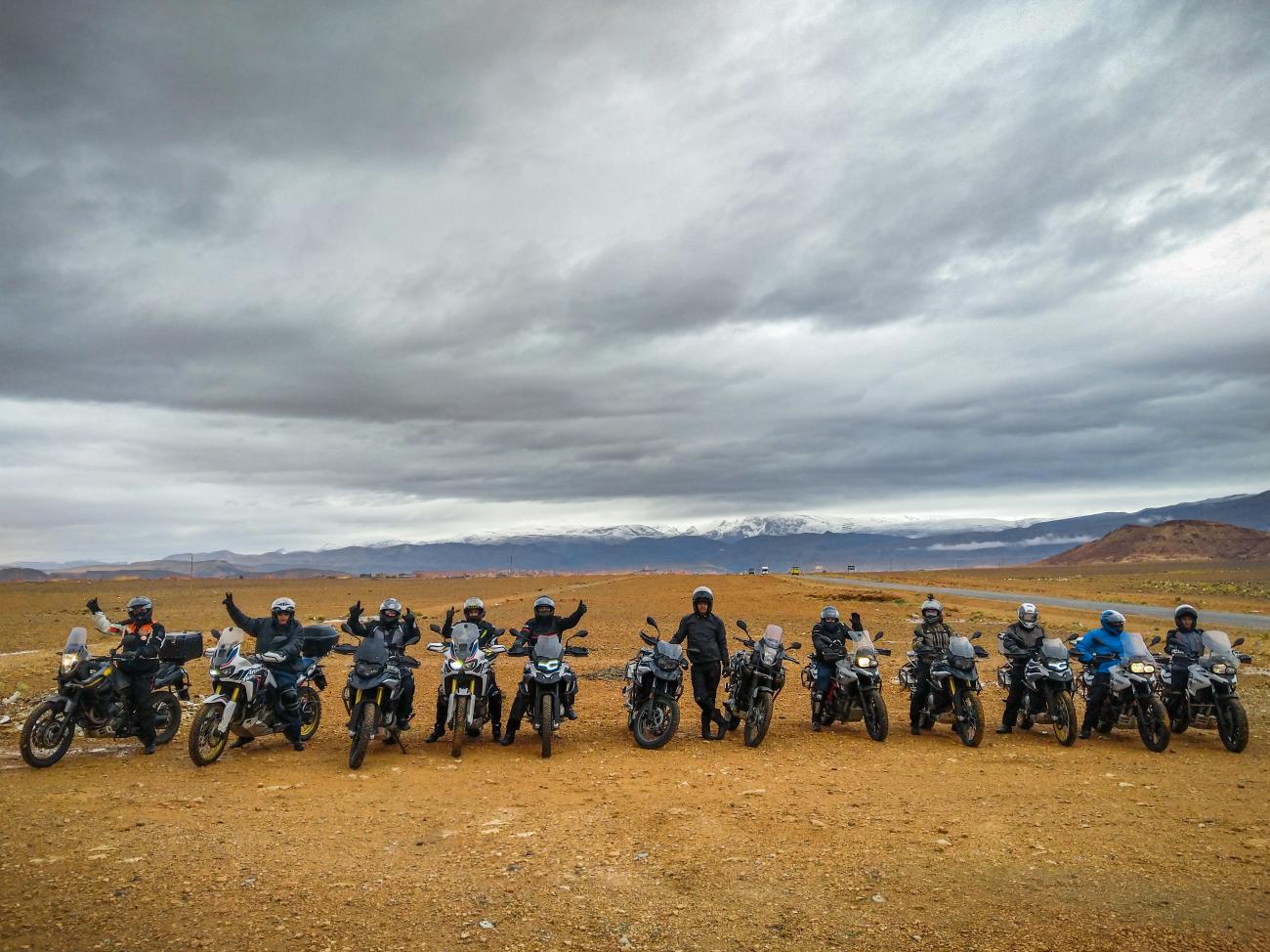 A large group of riders lined up with motorcycles on a plateau with snow-capped Atlas Mountains under dramatic clouds.