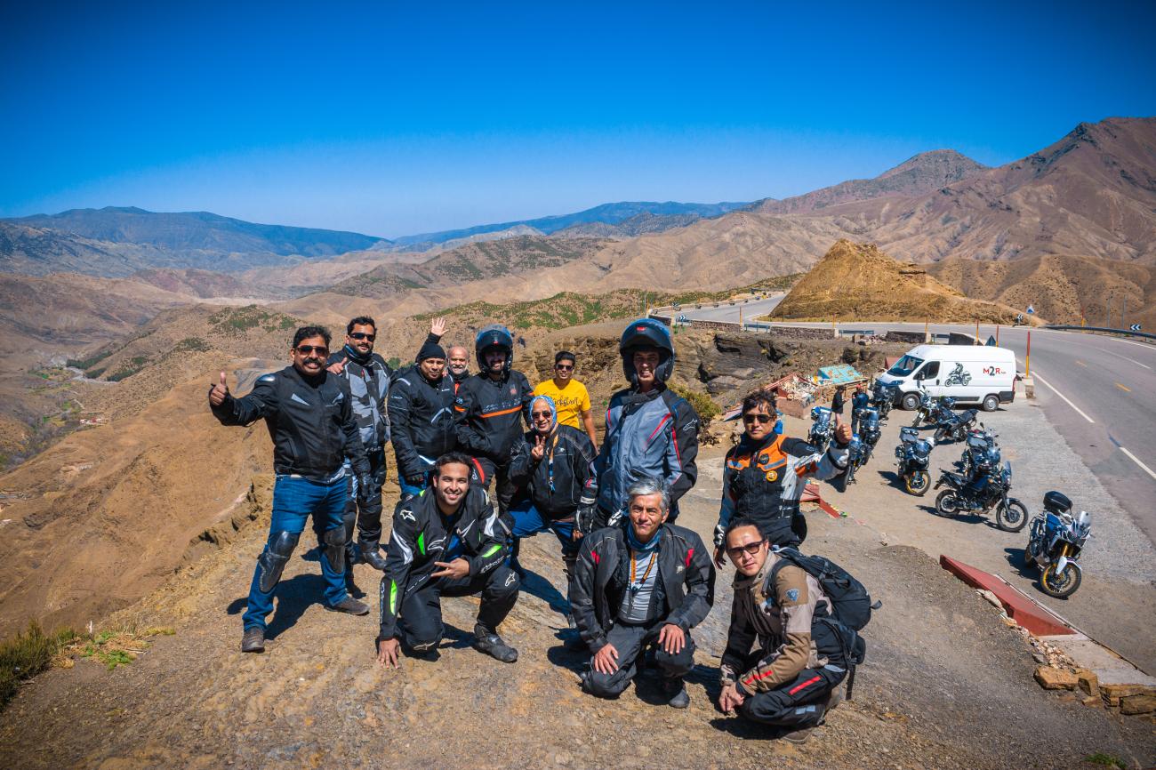 Motorcycle tour group posing at a high Atlas Mountain pass with sweeping mountain views in Morocco.