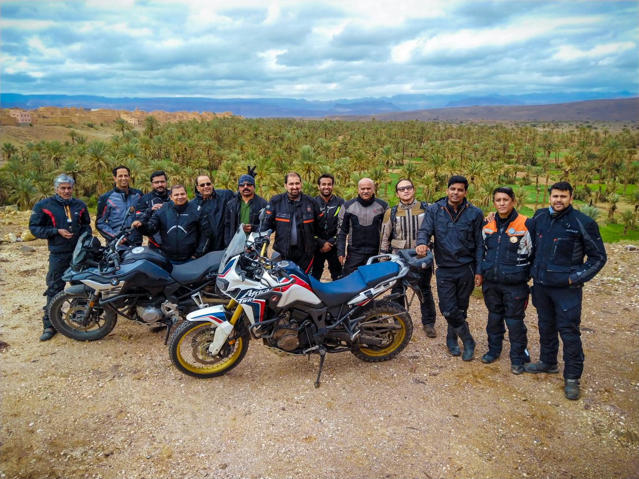 Motorcycle tour group posing in front of a lush palm grove oasis in Morocco.