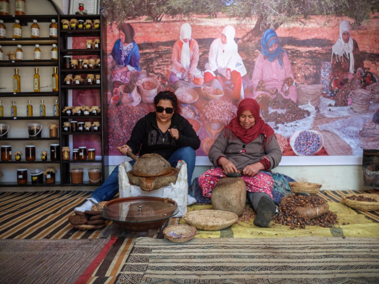 Traveller learning traditional argan oil preparation from a local Moroccan woman at a roadside workshop.