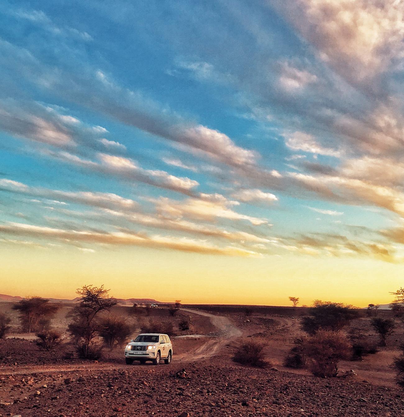 White SUV driving along a desert trail under a sweeping golden-hour sky in Morocco.