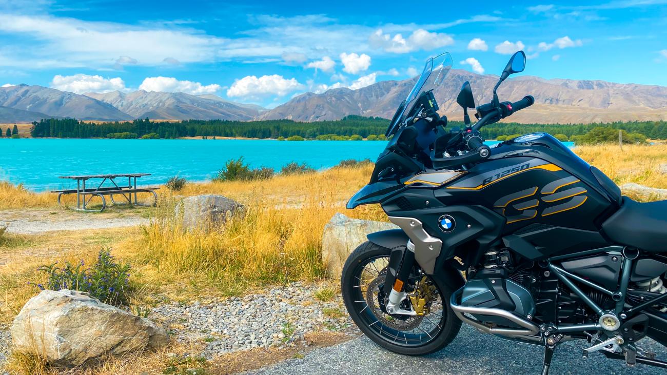 BMW GS adventure motorcycle parked at a lakeside rest area overlooking the turquoise waters of Lake Tekapo and surrounding mountains.
